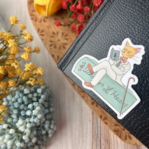May include: A sticker featuring a cartoon cat in a white suit, reading a book while reclining on a green surface. The sticker has the text "Wonder of the Heart" and a cane. The sticker is placed on a black book.