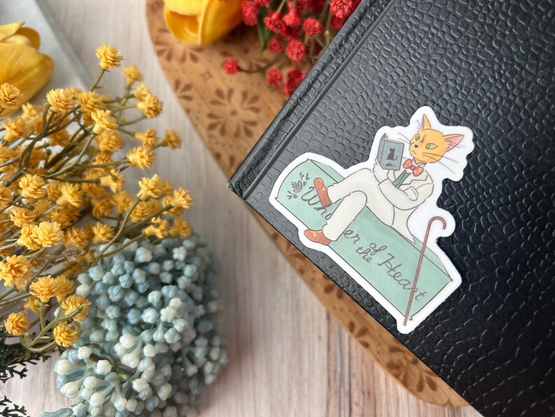 May include: A sticker featuring a cartoon cat in a white suit, reading a book while reclining on a green surface. The sticker has the text "Wonder of the Heart" and a cane. The sticker is placed on a black book.