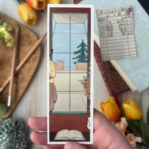 May include: A rectangular bookmark featuring an illustration of a window with a view of a tree and sky. The bookmark also depicts a person, a book, and a table. The color palette includes brown, blue, and beige.