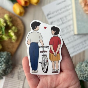 May include: A sticker featuring two cartoon figures, a boy and a girl, standing near a bicycle. The boy wears a white shirt and blue pants, while the girl wears a red shirt and yellow skirt. A red heart floats between them.