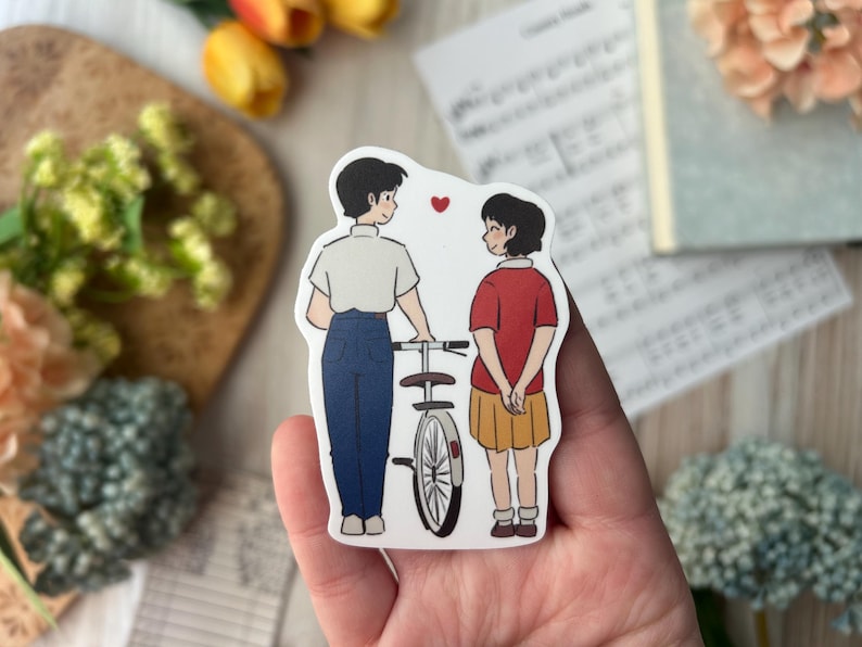 May include: A sticker featuring two cartoon figures, a boy and a girl, standing near a bicycle. The boy wears a white shirt and blue pants, while the girl wears a red shirt and yellow skirt. A red heart floats between them.