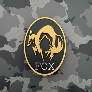 Puede incluir: Un parche redondo negro y dorado con una silueta de zorro estilizada y la palabra "FOX" en letras blancas.