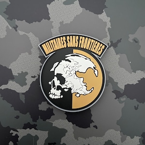Può includere: Un teschio bianco e nero su sfondo dorato. Il teschio è circondato da un cerchio nero con il testo "MILITAIRES SANS FRONTIERES" in bianco.