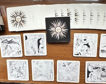 Op de afbeelding: Een set van 10 zwart-wit geillustreerde onderzetters met een zwarte doos. De onderzetters tonen verschillende scènes, waaronder een walvis, een brug en een zon. De doos heeft de tekst "The Sun & The Moon" erop gedrukt.