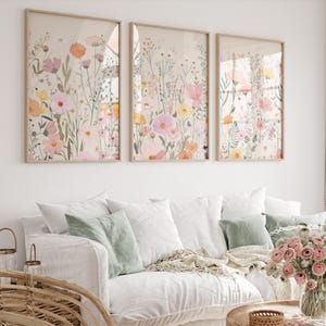 Puede incluir: Tres láminas enmarcadas con ilustraciones florales de acuarela de flores silvestres en tonos rosa, naranja, amarillo y verde. Las láminas se exhiben sobre un sofá blanco con cojines decorativos.
