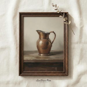 May include: A framed vintage print featuring a copper pitcher with a handle, set on a wooden surface. The artwork has a warm color palette with brown and beige tones. The frame is dark brown. The print is labeled "Luna Dream Prints".