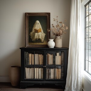 May include: A black wooden cabinet with glass doors filled with rows of books. On top sits a framed painting of a figure draped in a white sheet reading a book, a white ceramic vase, a tan textured vase, and a branch with dried flowers.