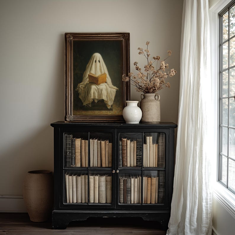 May include: A black wooden cabinet with glass doors filled with rows of books. On top sits a framed painting of a figure draped in a white sheet reading a book, a white ceramic vase, a tan textured vase, and a branch with dried flowers.