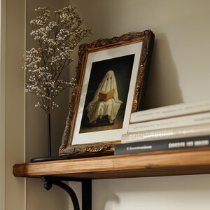 May include: A framed print featuring a figure draped in a white sheet reading a book, set within an ornate, dark gold-toned frame. The artwork is displayed alongside a dried baby's breath plant on a wooden shelf with a stack of books.