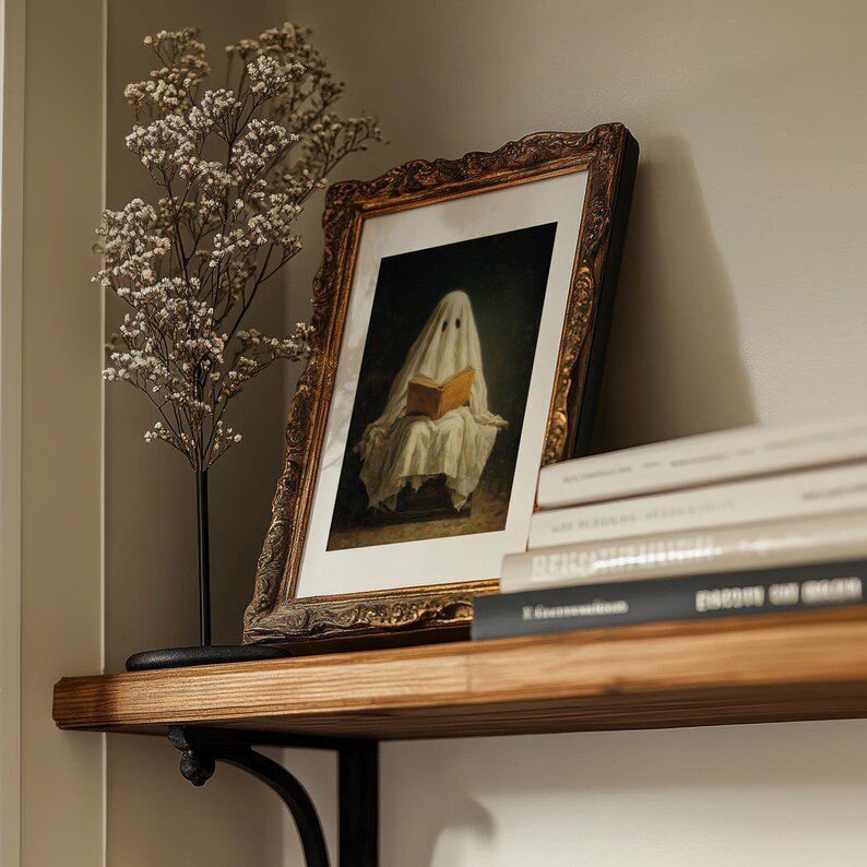 May include: A framed print featuring a figure draped in a white sheet reading a book, set within an ornate, dark gold-toned frame. The artwork is displayed alongside a dried baby's breath plant on a wooden shelf with a stack of books.