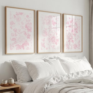 May include: Three rectangular wall art prints in light wood frames, each featuring a repeating pattern of soft pink floral illustrations on a white background, displayed in a horizontal row above a bed with white bedding and pillows.