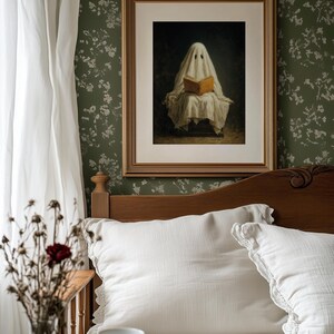 May include: A framed wall art print featuring a figure draped in a white sheet, resembling a ghost, sitting and reading an open book with a tan cover. The artwork is enclosed in a rectangular gold frame with a white mat, displayed on a dark green wall.