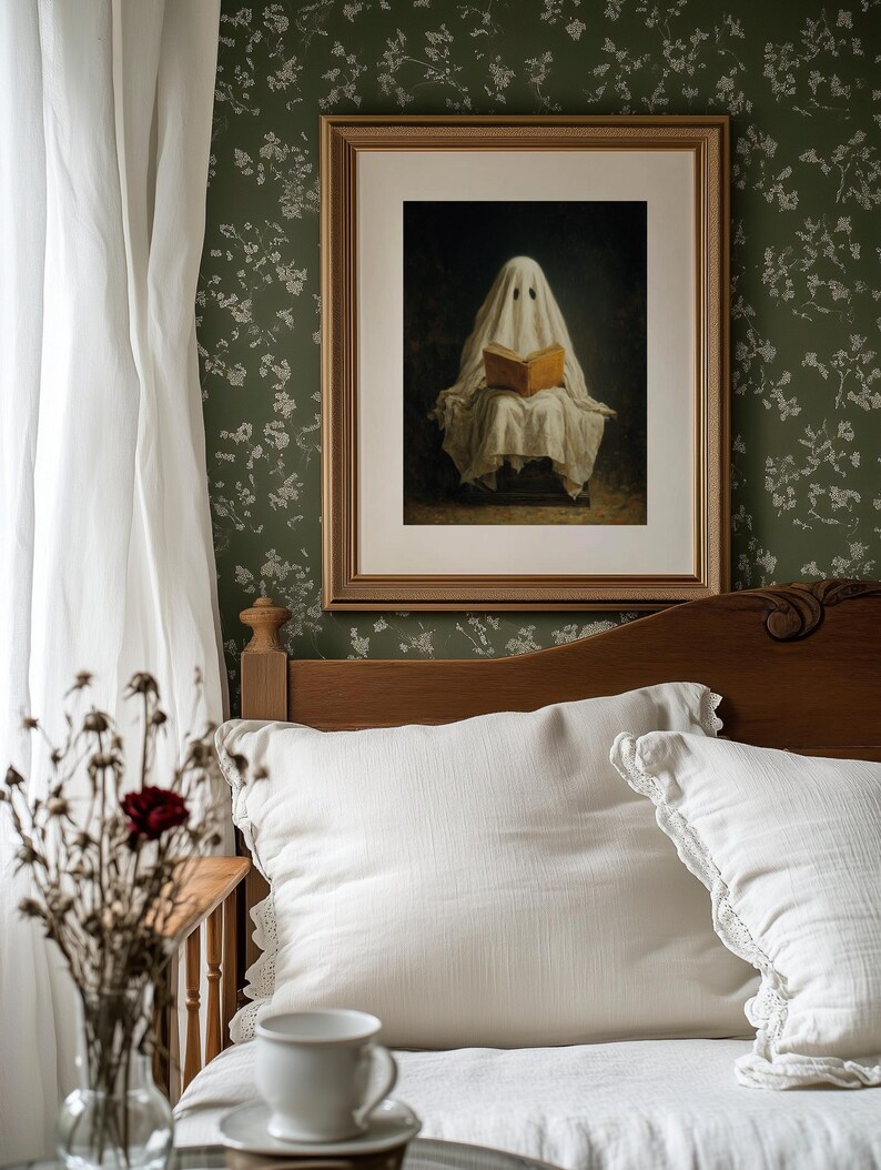May include: A framed wall art print featuring a figure draped in a white sheet, resembling a ghost, sitting and reading an open book with a tan cover. The artwork is enclosed in a rectangular gold frame with a white mat, displayed on a dark green wall.