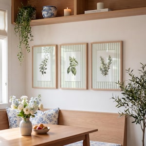 Op de afbeelding: Drie ingelijste botanische prenten met groene kruiden op een lichtgroene en wit gestreepte achtergrond. De prenten zitten in licht houten frames en hangen boven een houten tafel en bank. Een vaas met bloemen en een fruitschaal staan op de tafel.