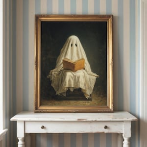 May include: A framed oil painting depicting a figure draped in a white sheet, resembling a ghost, sitting and reading an open tan book. The artwork is enclosed in an ornate gold frame, showcasing a dark, moody background behind the central figure.