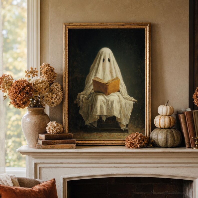 May include: A framed wall art print featuring a figure draped in a white sheet, resembling a ghost, sitting and reading an open book. The artwork is displayed on a mantelpiece alongside a ceramic vase with dried flowers and stacked pumpkins.