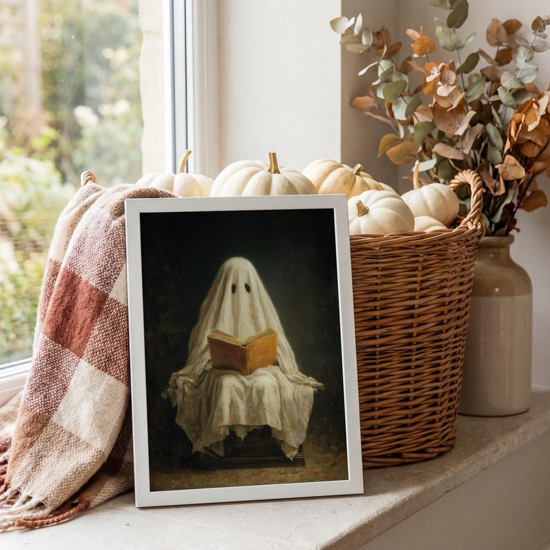 May include: A framed art print featuring a white ghost draped in a sheet, sitting and reading an open brown book against a dark background. The print is displayed in a white frame, positioned near a woven basket filled with white pumpkins.