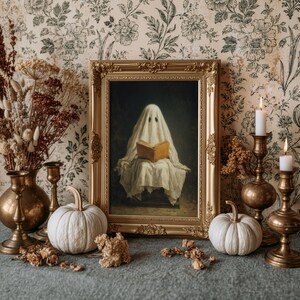 May include: A framed painting of a ghost draped in a white sheet reading an open book, displayed in an ornate gold frame. The scene includes two white pumpkins, two brass candlesticks with white candles, and a brass vase with dried flowers.