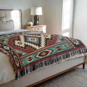 May include: A woven throw blanket with a Southwestern geometric pattern in shades of burgundy, teal, black, and cream. The blanket has a fringed edge and is draped over a bed. The design features diamond and zigzag shapes.