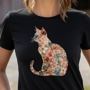 Può includere: Una maglietta nera con una stampa floreale di un gatto. Il gatto è seduto con la coda arrotolata attorno al suo corpo. I fiori sono in tonalità di rosa, rosso e blu.