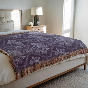 May include: A dark purple throw blanket with a floral rose pattern and fringed trim. The blanket is draped over a bed. The room has a neutral colour scheme. The bed has a neutral-coloured headboard and a wooden frame.