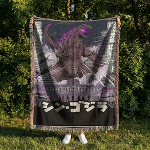May include: A black and purple woven throw blanket featuring a Godzilla design. The design includes the Japanese text "シン・ゴジラ" and other Japanese characters. The Godzilla illustration is in shades of purple and gray, with a building silhouette in the background.