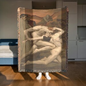 May include: A woven throw blanket featuring a classical painting of nude figures in a dramatic scene. The artwork has a warm color palette with figures in shades of beige and brown. The blanket has a fringed edge.