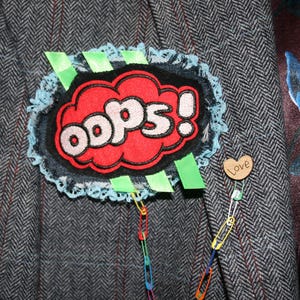 Pode incluir: Casaco de tweed cinzento com um patch decorativo que diz "oops!" em letras brancas dentro de uma nuvem vermelha. O patch é adornado com renda azul e fita verde. Também está preso um alfinete de madeira em forma de coração com a palavra "Love".