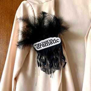 May include: A black feather and lace brooch with the text "a little bit dramatic" on a white background. The brooch is attached to a light pink blazer.