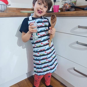 May include: A young child wearing a white and blue apron with a racing car pattern. The apron has a pocket on the front. The child is holding a wooden spoon and a glass of water. The child is standing in a kitchen.