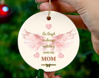 Op de afbeelding: Een crèmekleurige ronde kerstversiering met de tekst "An Angel is always watching over me MOM". De versiering heeft roze engelen vleugels en harten. Een gouden draad is bevestigd om op te hangen.