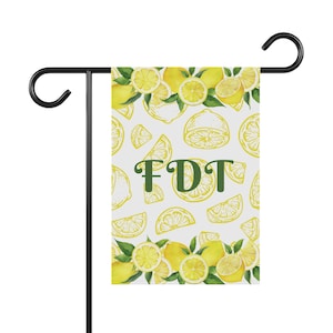 Könnte beinhalten: Eine weiße Gartenflagge mit einem Zitronenmuster und dem Text "FDT" in grünen Buchstaben. Die Flagge ist an einer schwarzen Metallstange befestigt.