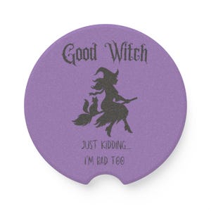 May include: A round, purple car coaster with a black silhouette of a witch on a broom with a cat. The text reads "Good Witch" above the image and "JUST KIDDING... I'M BAD TOO" below.