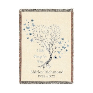 May include: Cream-colored woven throw blanket with a tree and butterfly design. The tree has butterflies floating away from the branches. Text reads "I Will Always Be Here" and "Shirley Richmond 1935-2022". The blanket has a fringed edge.