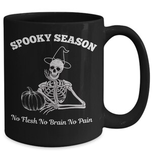May include: Black coffee mug with a white skeleton wearing a witch hat and holding a pumpkin. The text "Spooky Season" is above the skeleton and "No Flesh No Brain No Pain" is below.