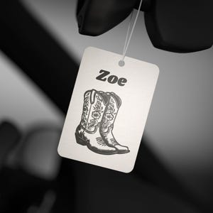May include: White car air freshener with a black and white illustration of cowboy boots and the name "Zoe" printed in black.