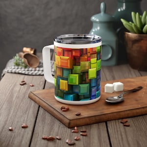 May include: A white travel mug with a colorful geometric design featuring squares in shades of red, orange, yellow, green, blue, and purple. The mug has a lid and a handle.