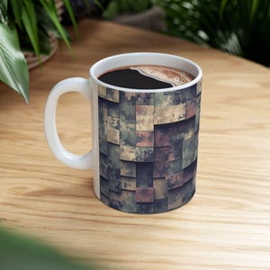 Könnte beinhalten: Weiße Keramik-Kaffeetasse mit einem geometrischen Muster in Grün-, Braun- und Grautönen. Die Tasse ist mit dunklem Kaffee gefüllt und steht auf einer Holzoberfläche. Der Griff ist weiß.
