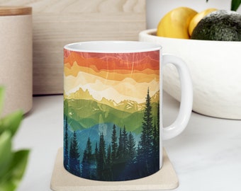 May include: White ceramic mug with a colorful mountain and forest design. The design features a rainbow gradient of red, orange, yellow, green, blue, and purple. The mountains are silhouetted against the gradient, and a forest of dark green trees is in the foreground.
