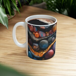 May include: White ceramic mug with a colorful abstract design of layered stones and swirls of orange, blue, and red. The mug is filled with coffee.