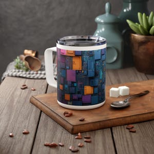 Könnte beinhalten: Weiße Keramiktasse mit einem farbenfrohen abstrakten Design aus Quadraten und Rechtecken in Blautönen, Lila, Orange und Rosa. Die Tasse hat einen Henkel und einen Deckel.
