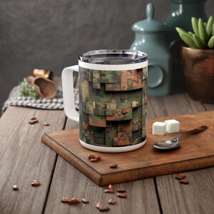 Könnte beinhalten: Eine weiße Keramiktasse mit Henkel, die ein Tarnmuster in Grün-, Braun- und Orangetönen aufweist. Die Tasse steht auf einer Holzoberfläche.