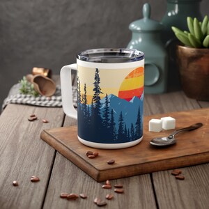 May include: White ceramic travel mug with a colorful sunset and mountain scene. The mug has a black handle and a lid.