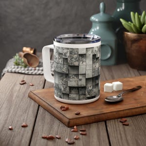Könnte beinhalten: Eine weiße Keramiktasse mit Henkel, die ein graues und weißes Tarnmuster aufweist. Die Tasse steht auf einer Holzoberfläche.