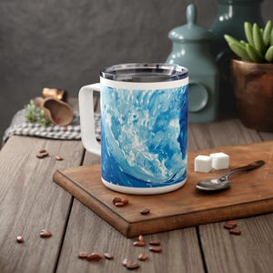 Könnte beinhalten: Eine weiße Keramiktasse mit einem abstrakten Wellenmuster in Blau und Weiß. Die Tasse hat einen Henkel und einen Deckel.