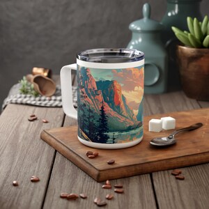 May include: White ceramic travel mug with a colorful mountain scene and lake. The scene features a mountain range with a lake in the foreground. The sky is a gradient of orange, pink, and blue.