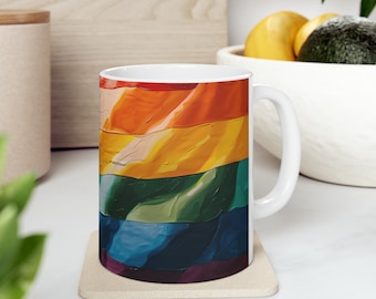 May include: White ceramic mug with a colorful rainbow flag design. The mug has a handle and is sitting on a beige coaster.
