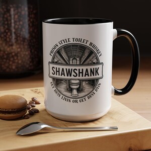 Puede incluir: Taza de cerámica blanca con interior y asa negros. La taza presenta el texto "PRISON STYLE TOILET WHISKEY SHAWSHANK GET BUSY LIVIN OR GET BUSY DYIN" en un diseño gráfico negro. Una cuchara y un macaron están sobre la superficie de madera.