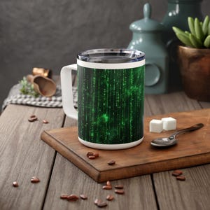 Op de afbeelding: Reismok met witte handgreep en een groen digitaal regenpatroon. De mok staat op een houten snijplank met suikerklontjes en een lepel. Koffiebonen zijn verspreid over de houten tafel.