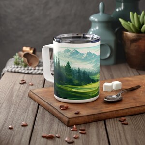 May include: White travel mug with a green, blue, and white landscape design featuring a golf course and mountains. The mug has a lid.